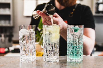 A person pours liquid into one of three glasses filled with ice cubes on a bar counter