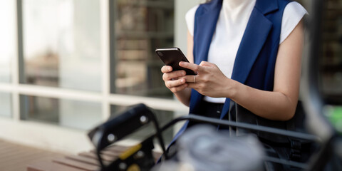 Businesswoman using smartphone while commuting by bicycle outside office. Concept of modern communication, connectivity, and sustainable transport