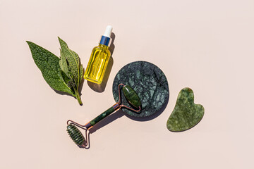 Natural cosmetic massage oil, roller massager and natural jade scraper on a beige background. top...