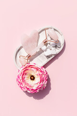 Rose quartz roller massager and gua sha scraper on a white ceramic tray with a delicate pink...