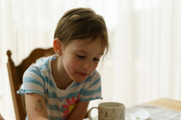 A small beautiful girl i eats a cupcake while sitting at a table against the backdrop of a large...