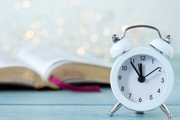 Alarm clock and open holy bible book with white bokeh light background. Close-up. Copy space. Christian biblical concept of patience, time, prayer, and Scripture study.
