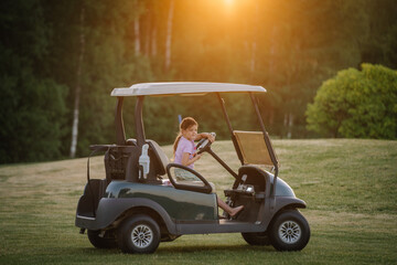 A young girl drives a golf cart on a grassy field at sunset, with a backdrop of lush green trees and a warm, golden glow.