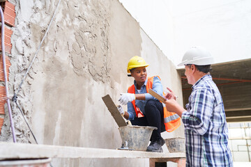 Senior male builder gesturing while talking to female mason plastering wall at construction site