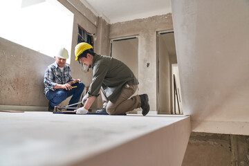 Full length of senior contractor crouching near worker cutting plank with equipment in corridor