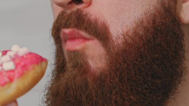 A bearded man is seen up close, enjoying a pink frosted donut with sprinkles and white cubes, savoring every bite