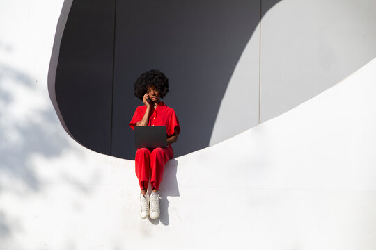 Mature woman in red jumpsuit with laptop talking through smart phone sitting on white wall - Powered by Adobe