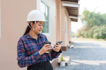 Asian female inspector in safety helmet holding coffee cup and smartphone. Depicts casual moment...