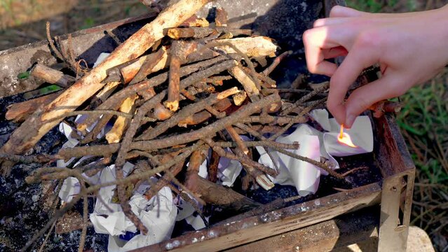 Young man sets fire to paper with match under clump of wooden branches for sawing grill. Holidays and active rest concept
