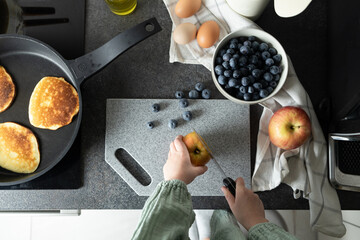 Child hands holding knife and cutting apple on stone cutting board. Preparation of pancakes on griddle