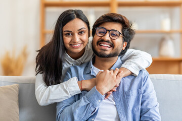 A joyful Indian couple embracing and smiling while sitting on a comfortable sofa in a warmly lit...