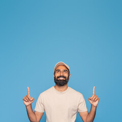 Indian man with a beard is smiling and pointing upwards with both hands. He is wearing a beige shirt and cap, standing in front of a bright blue background, copy space above