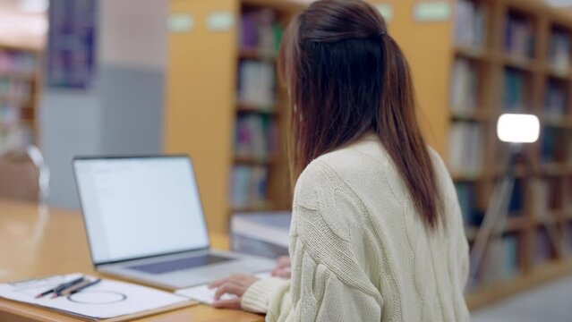 A woman is sitting at a desk with a laptop open in front of her. She is wearing a white sweater and has long brown hair. The scene suggests a quiet and focused atmosphere