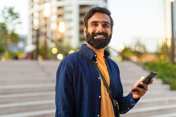 Indian man stands in an urban park, smiling as he holds his smartphone. He appears cheerful and...