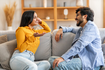 Indian man and woman are seated on a beige couch, engaged in conversation. Both individuals are facing each other, with the woman gesturing with her hands as they talk.