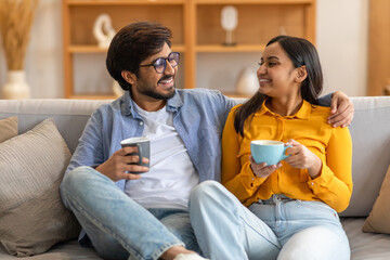 Indian man and woman are seated on a couch, each holding a cup of coffee. They appear relaxed as...