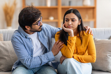 Indian young man and woman sit closely on a comfortable living room couch. The man is gently...