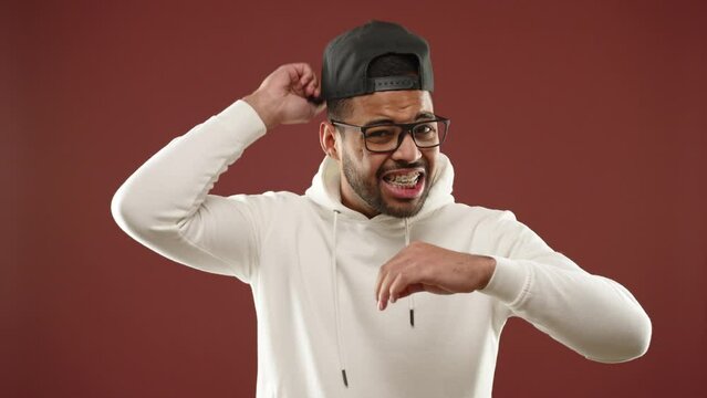 An exhausted African American man in a white hoodie is desperately wiping off profuse sweat from his forehead due to extreme heat, standing against a maroon backdrop. Camera 8K RAW. 