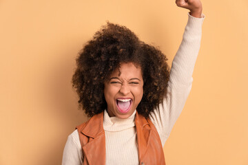 excited african american woman raising arms and celebrating in beige studio background....