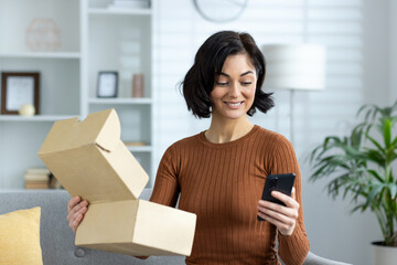 A smiling young woman is at home, sitting on the sofa, holding an open box of a parcel she received and looking at a mobile phone