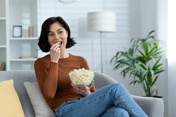 Young smiling girl resting at home. Sitting on the sofa, holding a bowl of popcorn and eating snacks