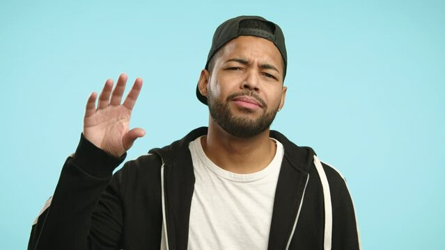 A communicative man in a cap with braces suggests there's too much gossiping going on by making a talking hand gesture on a light blue background. Camera 8K RAW. 