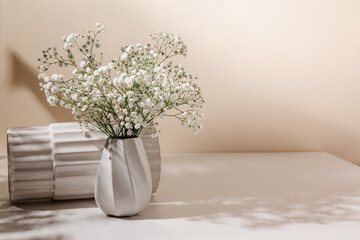 Beautiful still life composition with sunlight shadows. White ceramic vase with gypsophila flowers bouquet on the table with copy space, cozy interior for design or print