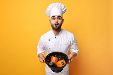 Surprised chef holding colander with vegetables on yellow background