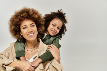 Stylish African American mother with curly afro hair carries her daughter on her shoulders against...