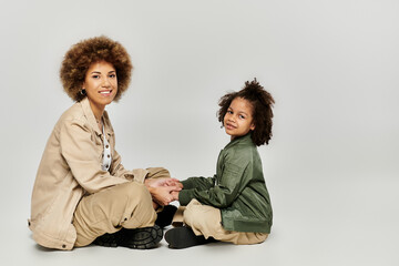Curly African American mother and daughter sitting together on grey floor in stylish clothes,...