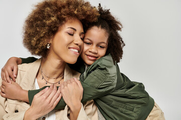 Curly African American mother and daughter in stylish clothes hugging fiercely in front of a white...