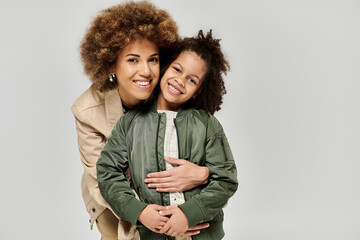 Curly African American mother and daughter in stylish clothes sharing a warm hug against a gray...