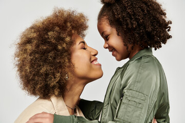 An African American mother and daughter, both with curly hair, sharing a loving hug against a gray...