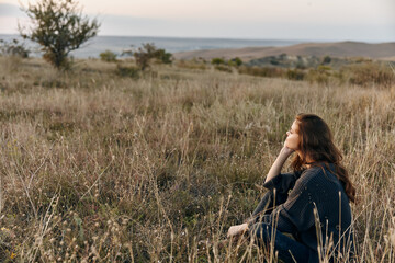 Woman sitting in peaceful grassy field, gazing up at the endless blue sky filled with fluffy white...