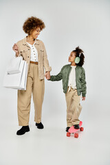 Fototapeta premium Curly African American mother and daughter in stylish clothes hold shopping bags and hands against a white background.