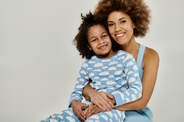 Joyful African American mother and daughter hugging tightly in cozy pajamas on a grey background.