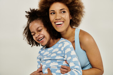 A happy African American mother wearing pajamas hugging her daughter tenderly in front of a white...