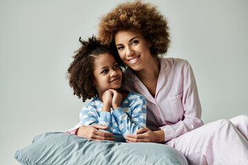 An African American mother and daughter in pajamas happily posing for a photo on a grey background.