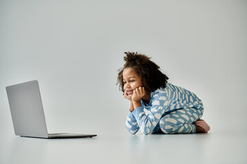 A happy African American girl in pajamas sits on the floor engrossed in a laptop