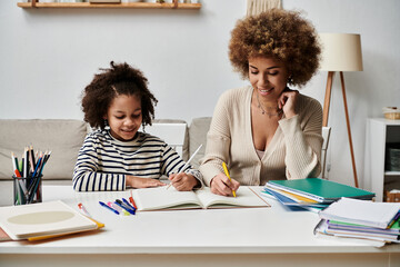 An African American mother and her daughter sit at a table, engrossed in doing homework together,...