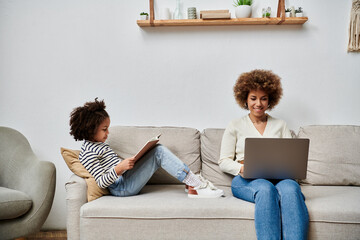 An African American mother and daughter happily seated on a couch, engrossed in using a laptop...