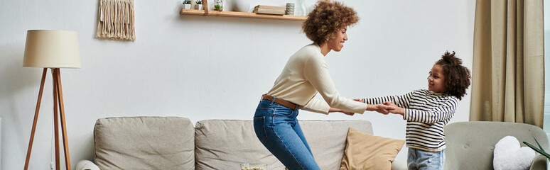 An African American mother and daughter gleefully dance together on a couch, enjoying quality time...
