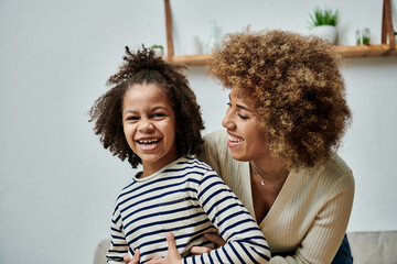 A happy African American mother and daughter are laughing together while sitting on the couch in...