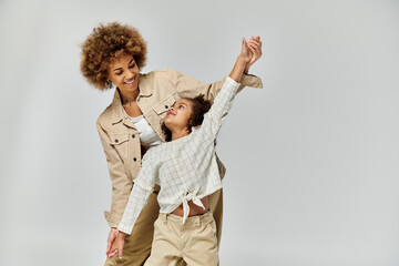 A curly African American mother and daughter joyfully dancing in stylish outfits on a gray background.