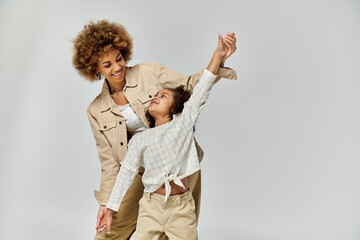A curly African American mother and daughter joyfully dancing in stylish outfits on a gray...
