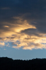 Landscape of thick clouds, orange light hitting the blue sky, silhouettes of trees, forests, and mountains. Area for text in the middle of the image
