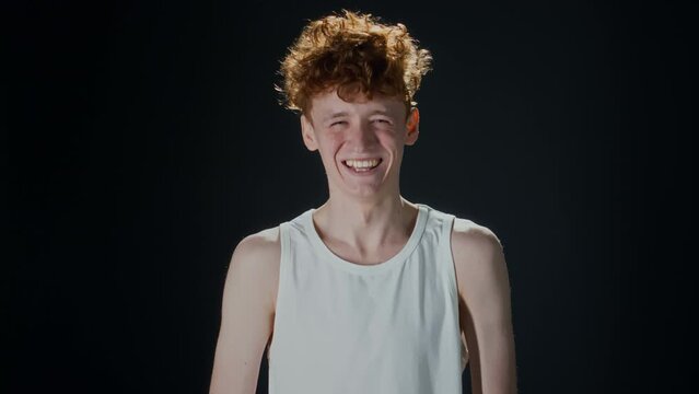Studio portrait of gen Z man in undershirt and with red curly hair looking at camera and laughing non stop against black background