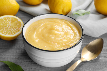 Delicious lemon curd in bowl, fresh citrus fruits and spoon on grey wooden table, closeup