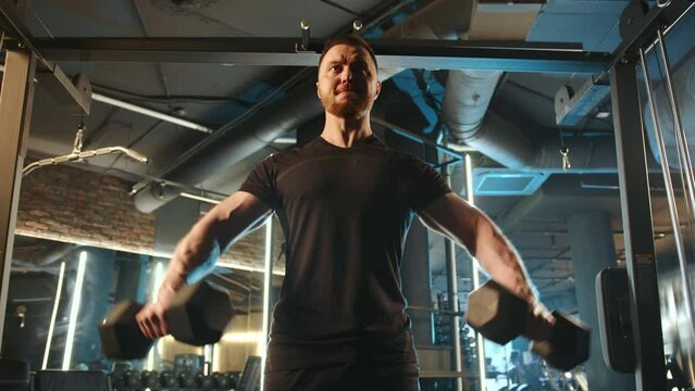 A determined man doing dumbbell lateral raises at the gym. Strength, focus, and fitness dedication are evident as he works on his shoulder muscles in a well-equipped gym environment.