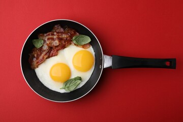 Fried eggs, bacon and basil in frying pan on red background, top view